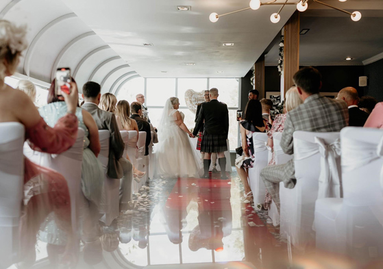 Bride and groom stand at altar holding hands during ceremony, while guests look on from white chairs.
